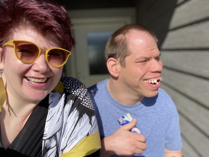a neurodiverse man sits next to a woman wearing sunglasses. Both have big smiles.